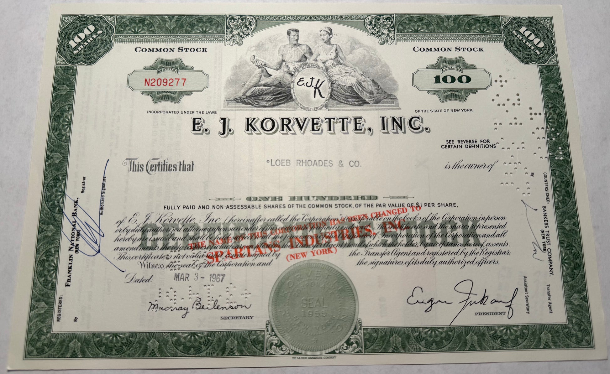 This green stock certificate from E.J. Korvette, issued in 1967, features cancellation holes, stamps, and handwritten annotations. The certificate also bears a mark indicating the company's name change to "Spartans Industries."